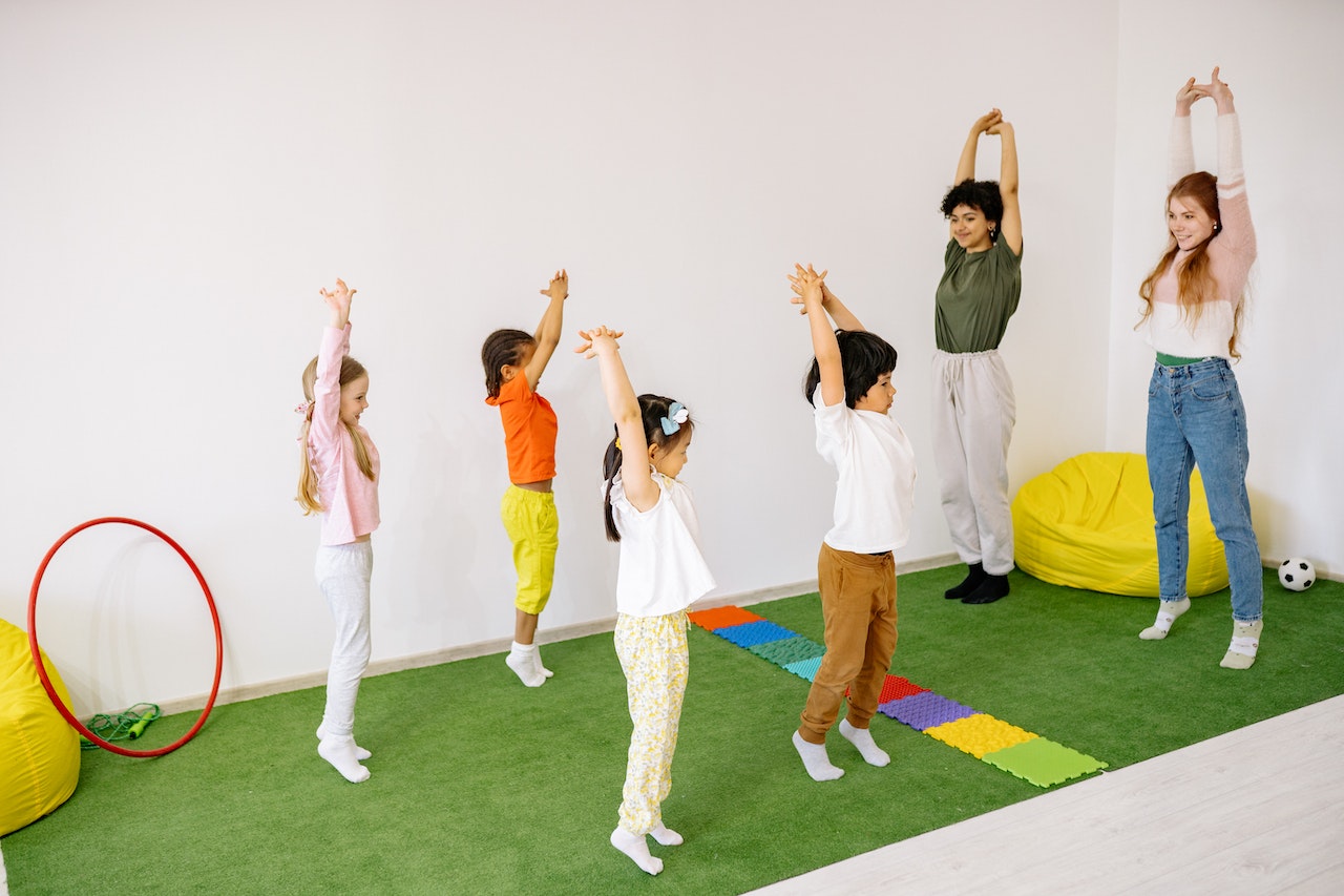 Dos instructoras guían a cuatro niños en estiramientos hacia arriba en una clase de yoga para niños en una sala de juegos